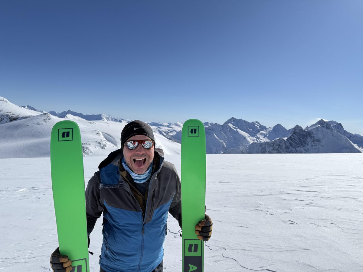 Jean-Sébastien Fontaine ski touring, holding a pair of skis on a snowfield below a range of peaks